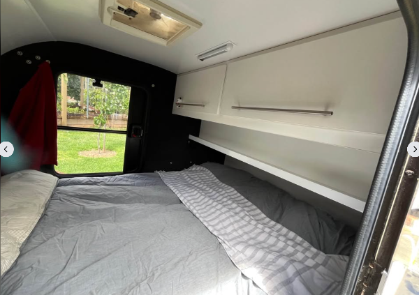 A Little Guiy camper interior showing a bed with gray bedding, white overhead storage cabinets, and an open rear door revealing acomplete camping set up with My Tesla Accessories
