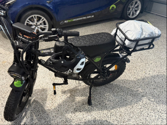 Black Ouxi V8 electric e-bike with a cargo basket on a gravel surface, with a blue car in the background.